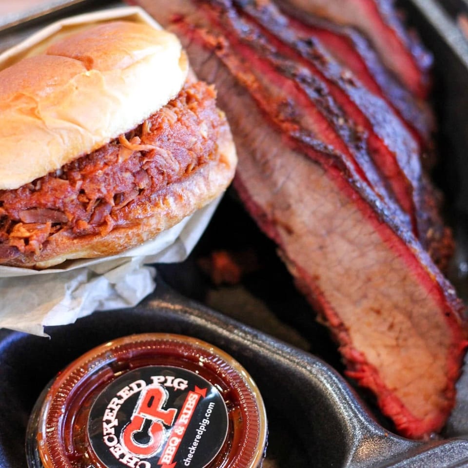 A Checkered Pig BBQ & Ribs pulled pork sandwich, sliced brisket, and sauce in a to-go container.