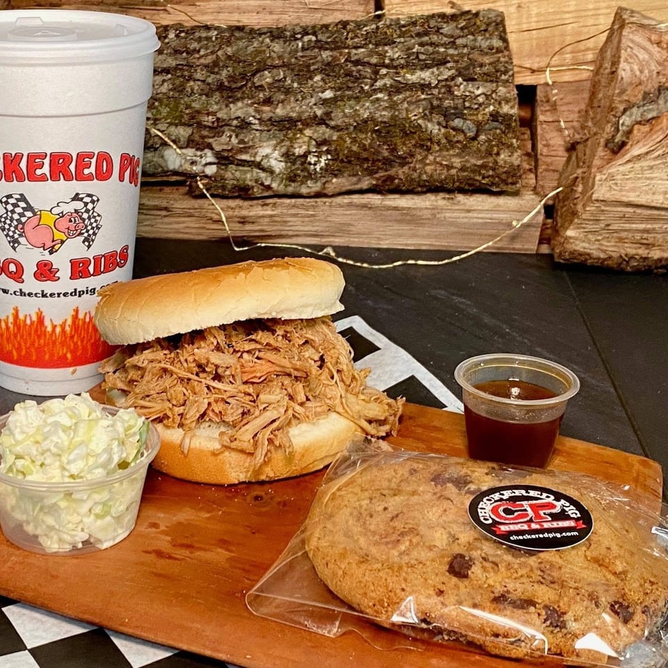 Checkered Pig BBQ pulled pork sandwich, coleslaw, sauce, drink, and a chocolate chip cookie on a wooden board.