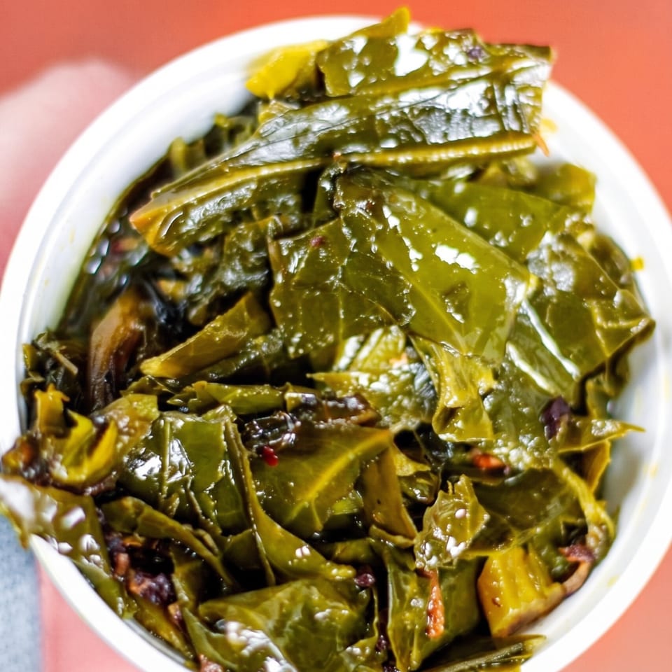 A bowl of glossy sautéed green vegetables with red chilies, served on a white plate against a coral background.