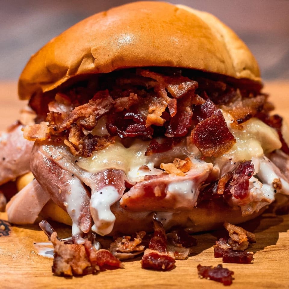 A close-up of a loaded sandwich with bacon, melted cheese, and sauce on a wooden surface.