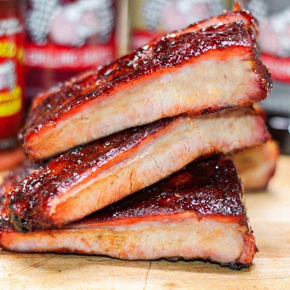 Three glazed barbecue ribs stacked on a wooden cutting board with sauce bottles in the background.
