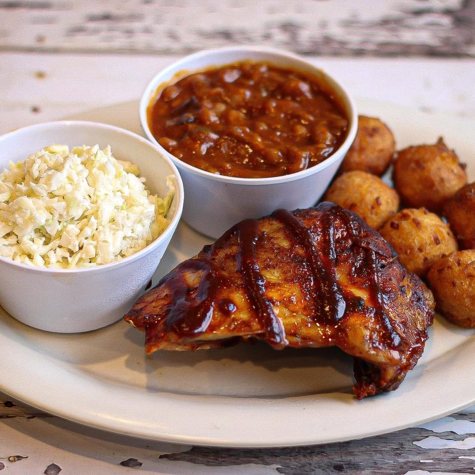 Barbecue chicken with baked beans, coleslaw, and hush puppies on a white plate.